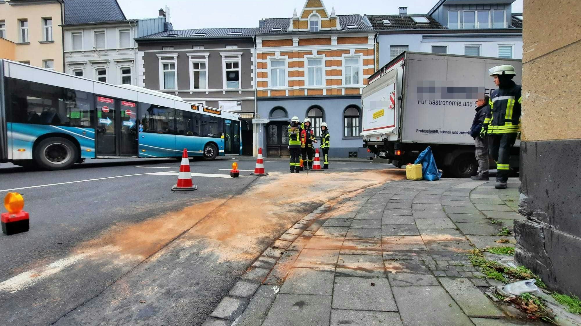Ein blau-weißer Bus passiert an der Unfallstelle vorbei die Ecke Linzer Straße/Am Saynschen Hof. Mit bräunlichen Bindemitteln haben Feuerwehrleute die ausgelaufenen Betriebsstoffe abgestreut.
