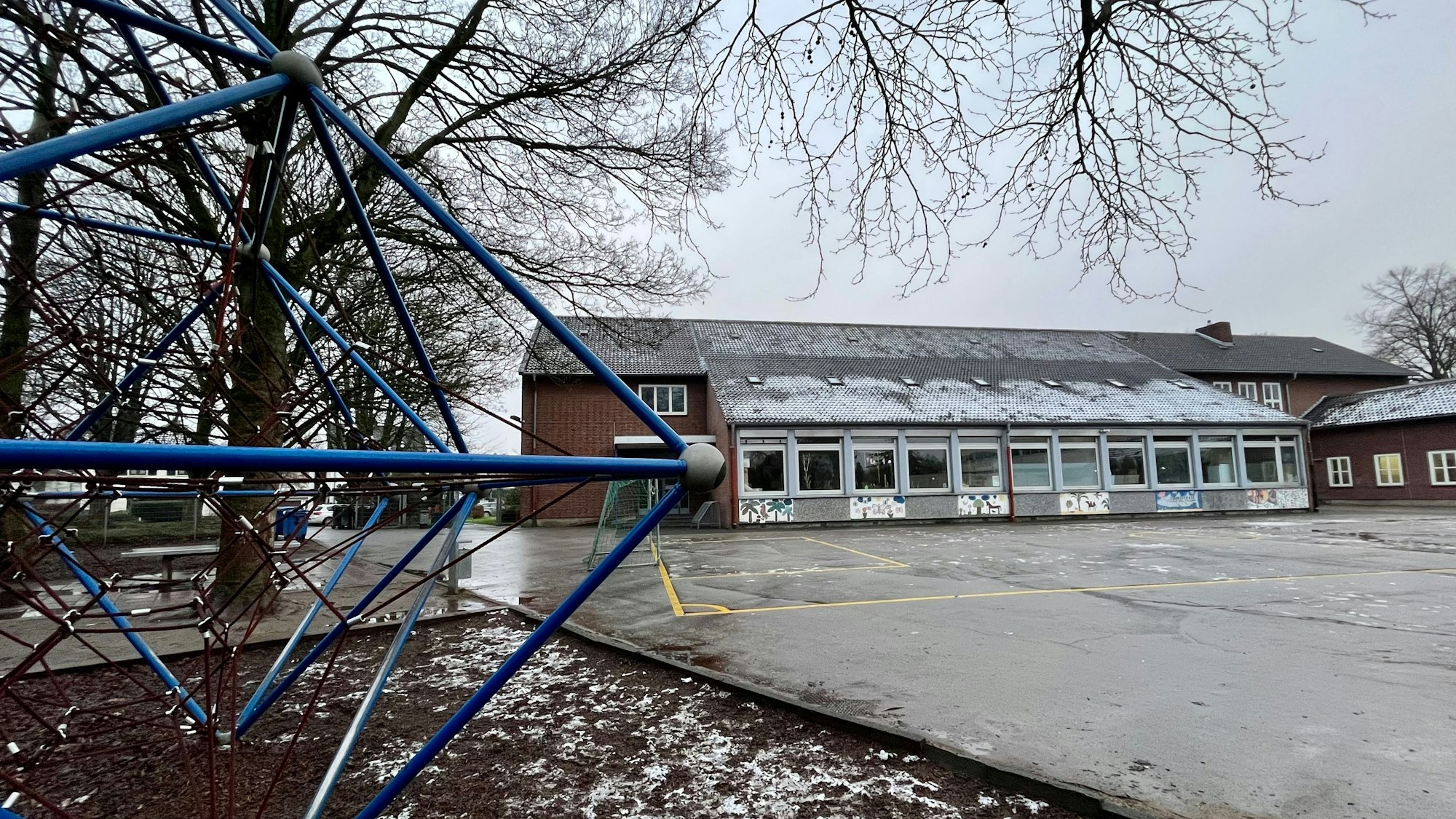 Ein Klettergerüst im Vordergrund, ein Schulhof und ein flaches Schulgebäude im Hintergrund.