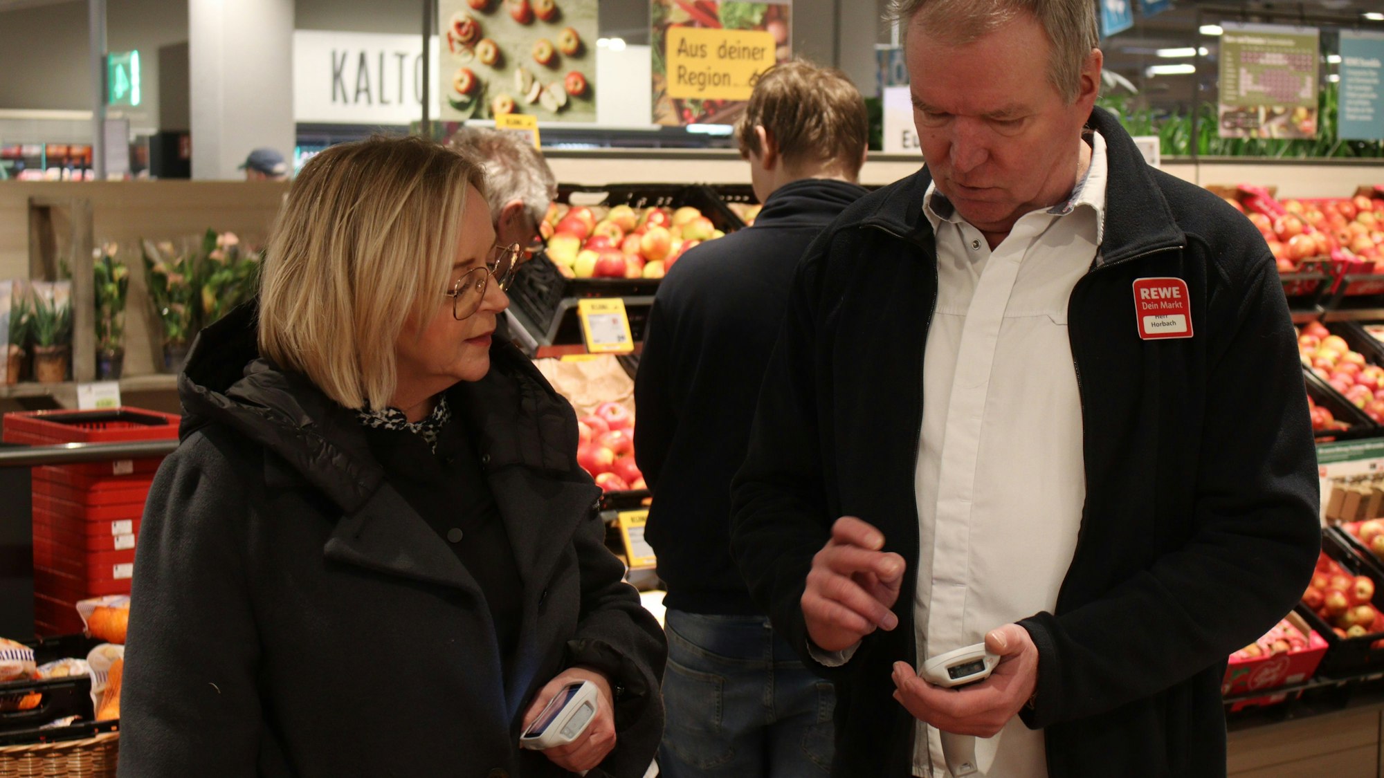 Marktmanager Achim Horbach zeigt Bürgermeisterin Sabine Preiser-Marian in der Obstabteilung des Rewe-Centers, wie die Warenscanner funktionieren, mit denen Kunden nun während des Einkaufs ihre Produkte scannen können.