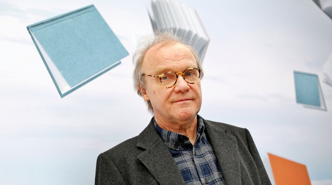 Der österreichische Autor Michael Köhlmeier steht in einem dunklen Jackett und einem karierten Hemd vor einer mit Büchern bedruckten Wand.