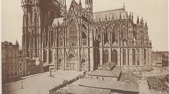 Das historische Foto vom Weiterbau des Kölner Doms zeigt den Zustand im Mai 1876 mit den noch unvollendeten Türmen. Im Vordergrund sieht man die Dombauhütte.