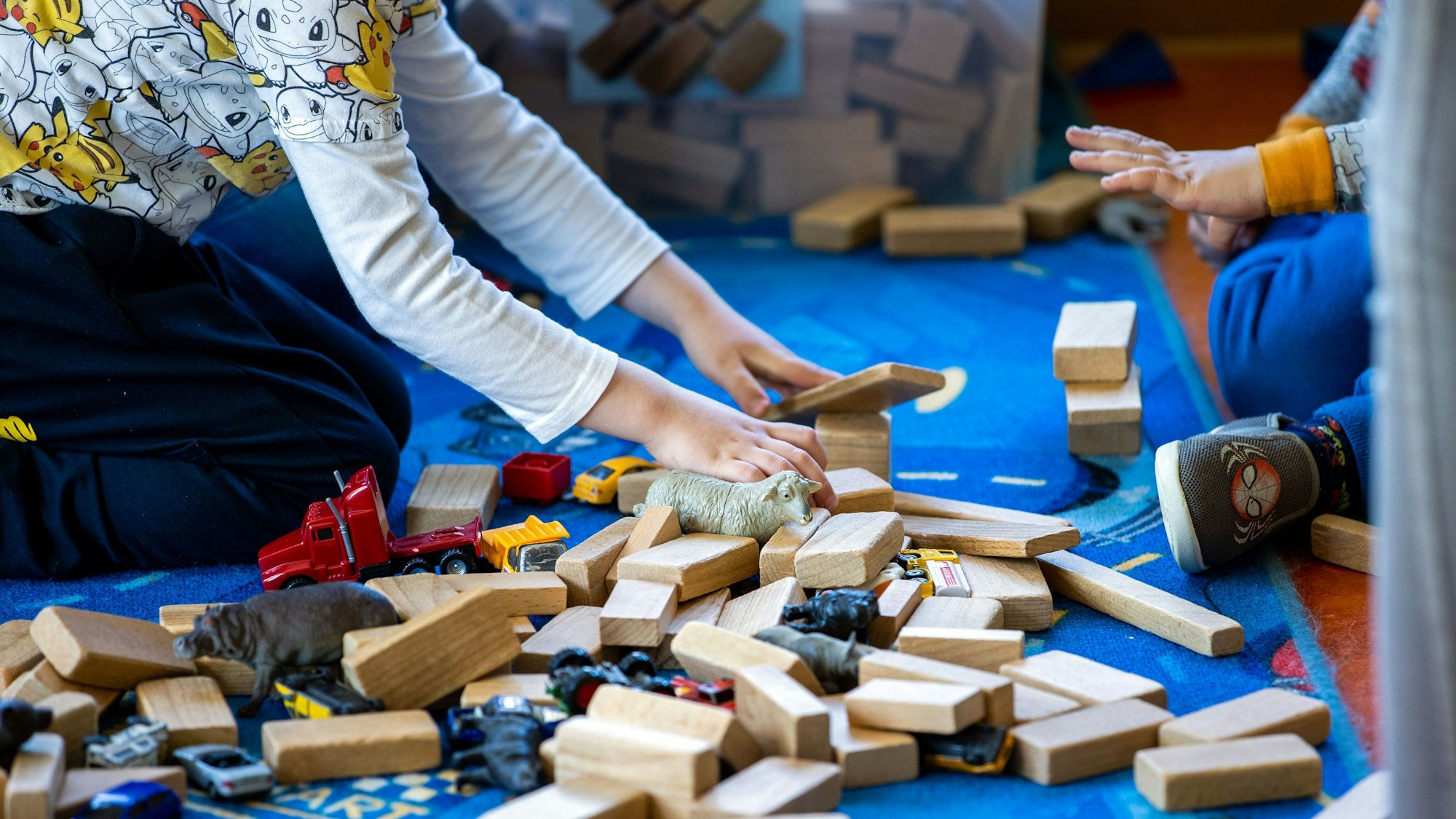 In einer Kindertagesstätte liegen Holzbauklötze, Spielzeugautos und Tierfiguren auf dem Fußboden, auf dem zwei Kinder spielen.