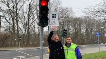 An einer roten Ampel stehen zwei Männer, einer hält ein Schild mit einem grünen Pfeil und dem Symbol eines Fahrrades in die Höhe.