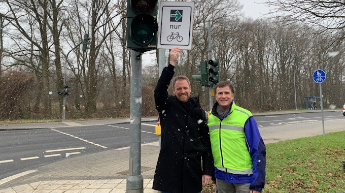 An einer roten Ampel stehen zwei Männer, einer hält ein Schild mit einem grünen Pfeil und dem Symbol eines Fahrrades in die Höhe.