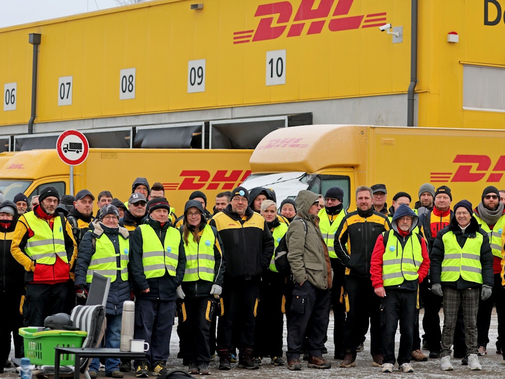 Mitarbeiterinnen und Mitarbeiter der Deutschen Post stehen in gelben Warnwesten vor dem Zustellstützpunkt. Hinter ihnen stehen die Postautos.