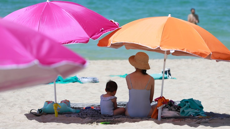 Eine Frau sitzt neben ihrem Kind unter einem Sonnenschirm an einem warmen Sommertag am Strand von Mallorca.