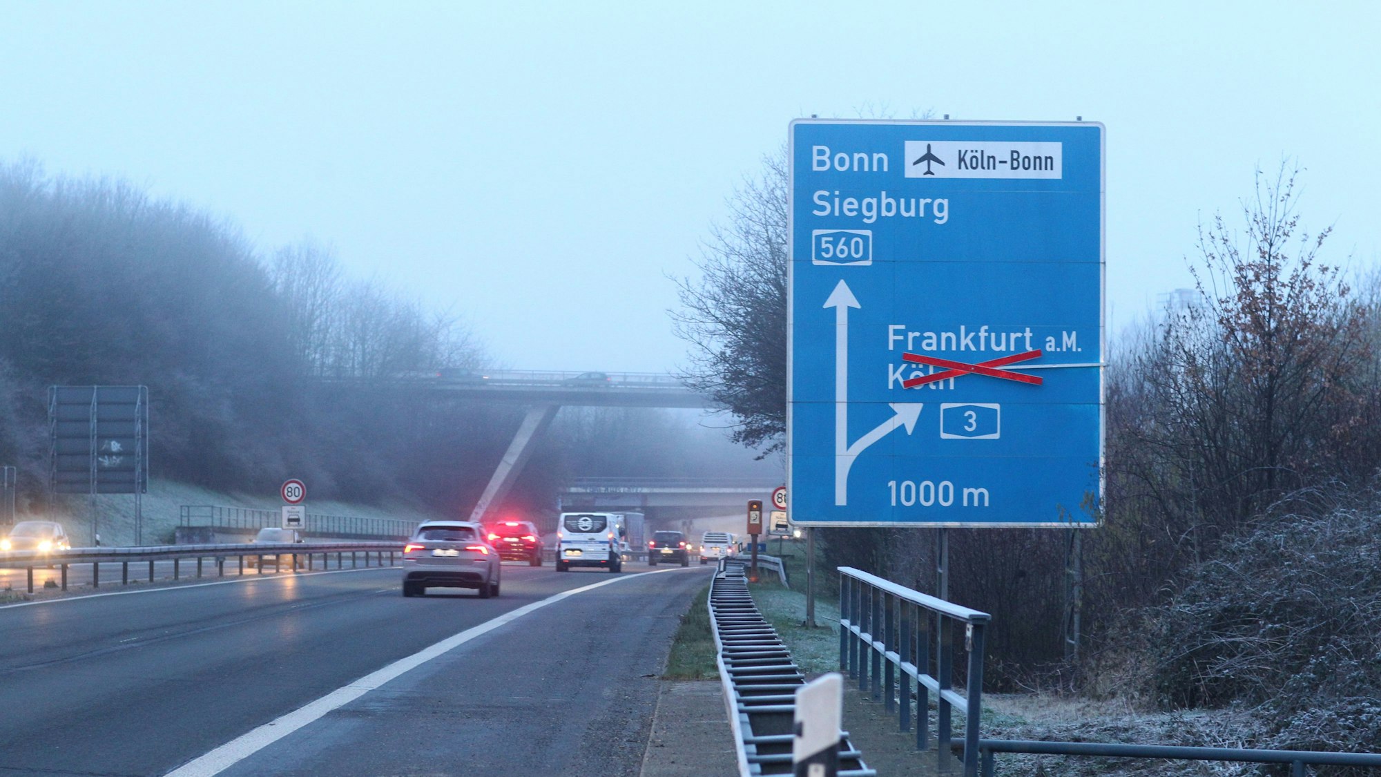 Autos bremsen auf einer winterlichen Autobahn, die Rücklichter leuchten. Im Vordergrund steht ein blaues Schild, das die Fahrspur der A560 Richtung Bonn und Siegburg anzeigt. Der Pfeil Richtung Köln ist mit einem Kreuz durchgestrichen.