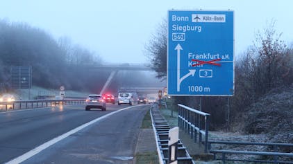 Autos bremsen auf einer winterlichen Autobahn, die Rücklichter leuchten. Im Vordergrund steht ein blaues Schild, das die Fahrspur der A560 Richtung Bonn und Siegburg anzeigt. Der Pfeil Richtung Köln ist mit einem Kreuz durchgestrichen.