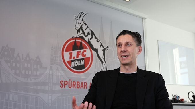 Christian Keller, Geschäftsführer des 1. FC Köln, gestikuliert vor einem FC-Plakat mit Geißbock