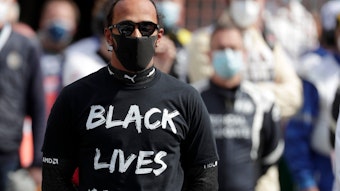 Man sieht Lewis Hamilton im Vordergrund mit schwarzem T-Shirt mit Aufdruck „Black Lives Matter“, schwarzer Maske und Sonnenbrille. Im Hintergrund unscharf weitere Menschen.