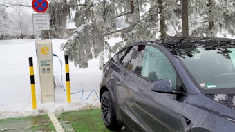 Ein Elektroauto steht an einer Ladestation, im Hintergrund ein Baum und eine Wiese, die mit Schnee bedeckt sind.