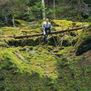 Ein Mann im Anzug sitzt mit einem Laptop auf einem umgestürzten Baumstamm im Wald.