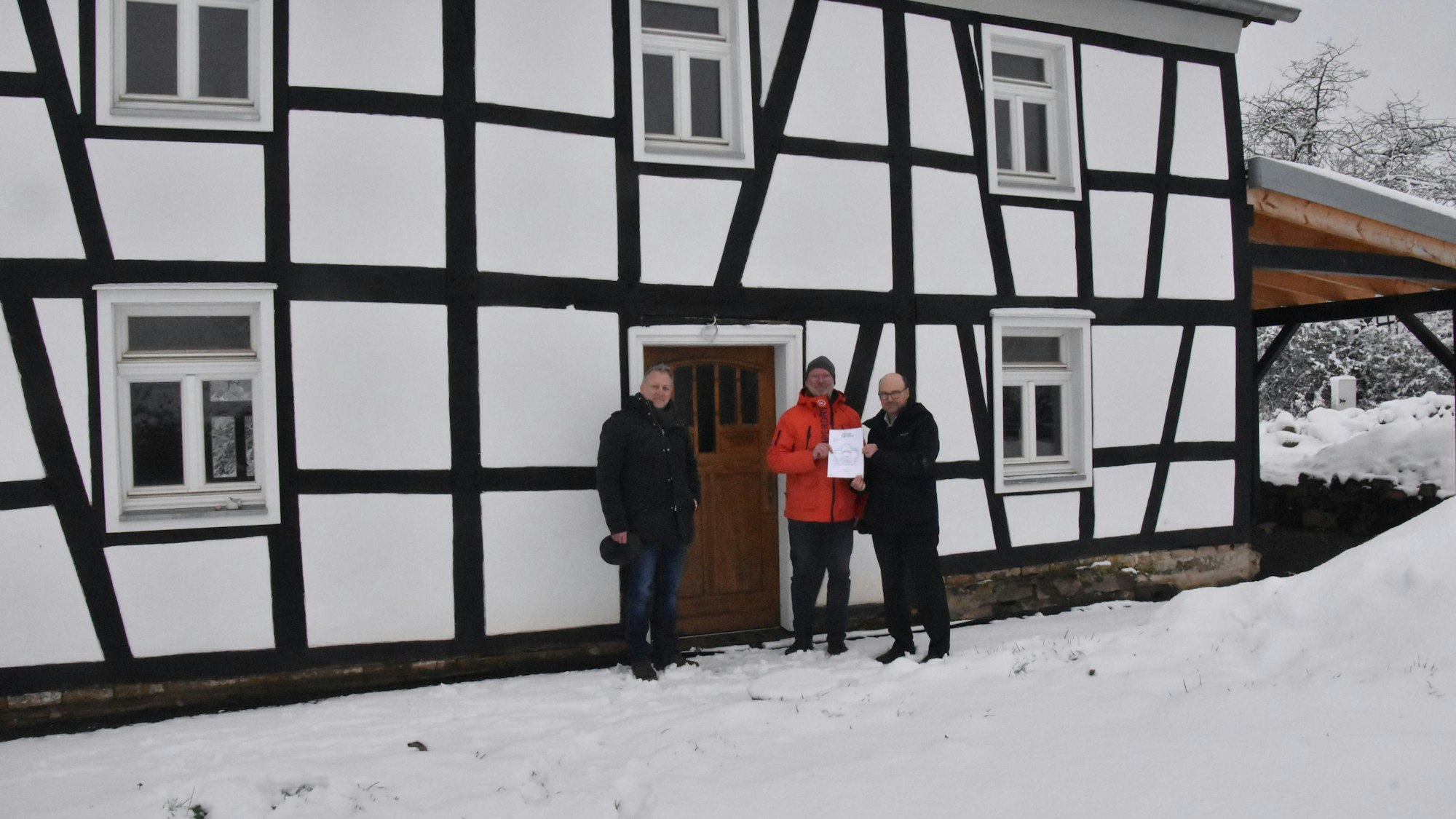 Drei Männer stehen im Schnee vor einem Fachwerkhaus, rechts und links neben der hölzernen Eingangstür. Zwei halten ein bedrucktes Blatt Papier in die Höhe.