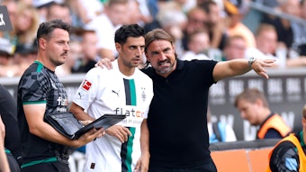 Daniel Farke (r.) legt seinen Arm um Lars Stindl (M.) und gibt dem Kapitän von Borussia Mönchengladbach vor einer Einwechslung taktische Anweisungen. Farke macht zudem eine Geste mit seinem linken Arm. Neben Farke und Stindl steht Co-Trainer Edmund Riemer und hält eine Mappe in der Hand.