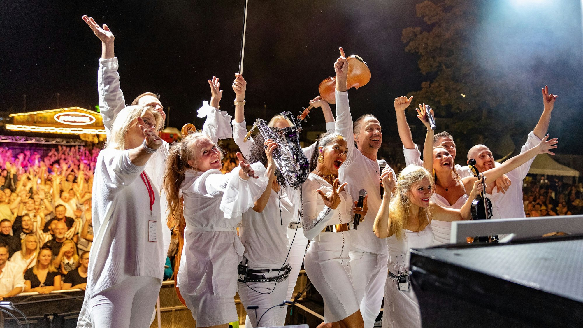 Menschen in weißer Kleidung, mit Instrumenten in der Hand, jubeln auf einer Bühne.