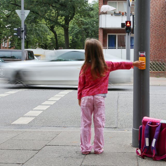 Ein rosa gekleidetes Mädchen steht mit Ranzen an einer Ampel, um die Straße zu überqueren.
