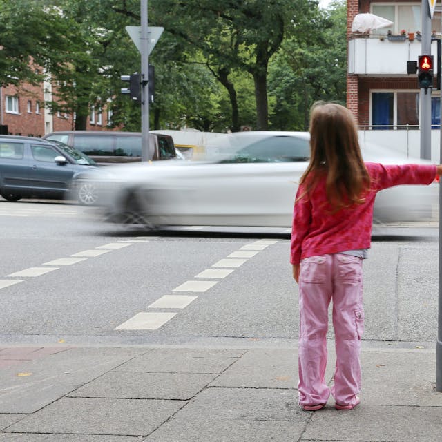 Ein rosa gekleidetes Mädchen steht mit Ranzen an einer Ampel, um die Straße zu überqueren.