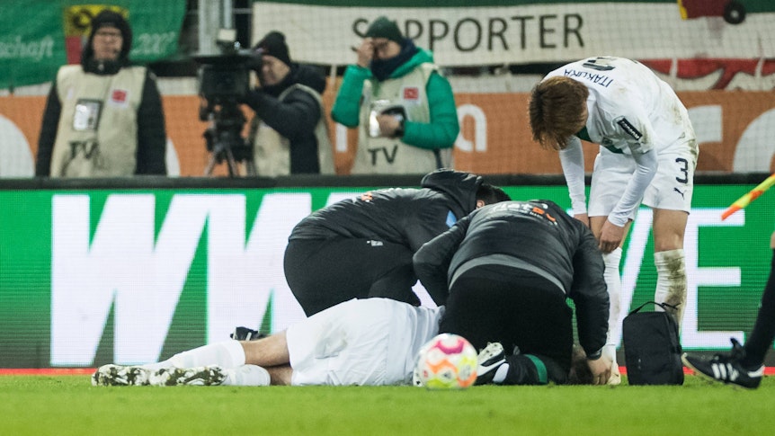 Christoph Kramer wird auf dem Rasen in der WWK-Arena in Augsburg behandelt. Die Mannschaftsärzte von Borussia Mönchengladbach und Mitspieler Ko Itakura stehen um ihn herum.