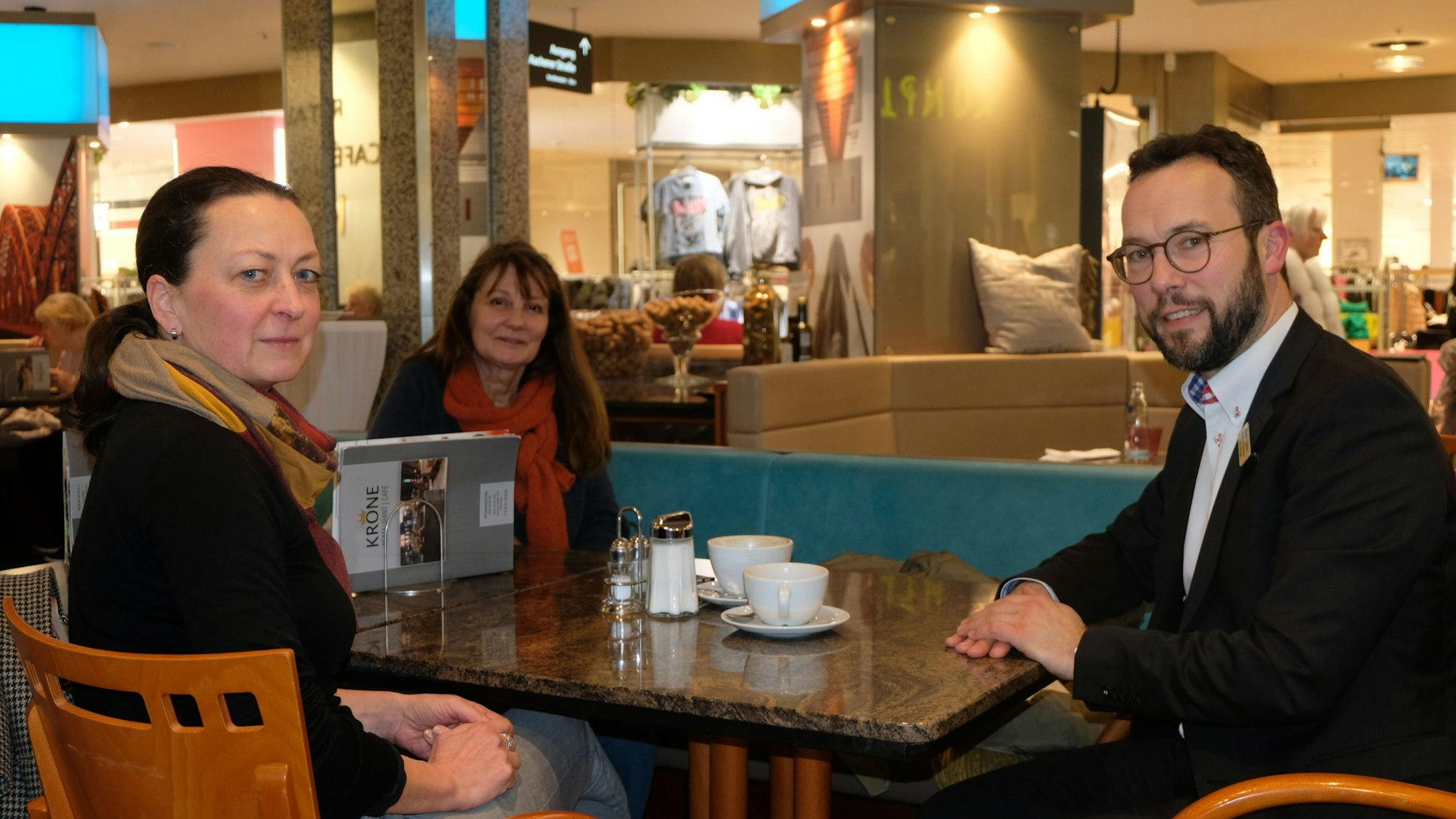 Zwei Frauen mit dunklen Haaren und ein Mann mit Bart und Brille sitzen an einem Café-Tisch im Einkaufszentrum Rhein-Center.