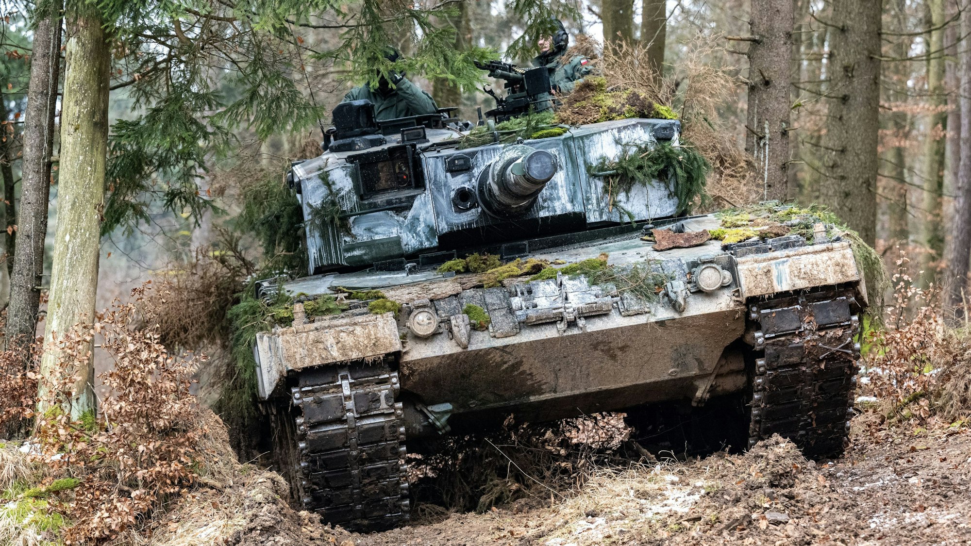 In einem Nadelwald bewegt sich ein zum Teil mit Moosen getarnter Panzer des Typs Leopard 2 zwischen Baumstämmen hindurch. Zwei bewaffnete Soldaten ragen aus den Panzereinstiegsöffnungen heraus.