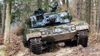In einem Nadelwald bewegt sich ein zum Teil mit Moosen getarnter Panzer des Typs Leopard 2 zwischen Baumstämmen hindurch. Zwei bewaffnete Soldaten ragen aus den Panzereinstiegsöffnungen heraus.