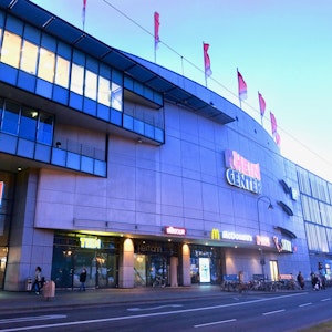 Ein mehrstöckiges Gebäude mit Fahnen auf dem Dach und dem Schriftzug Rhein-Center über Glas-Eingangstüren ist zu sehen.