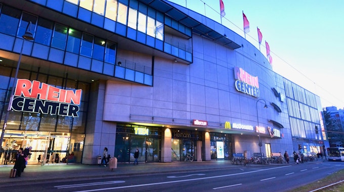 Ein mehrstöckiges Gebäude mit Fahnen auf dem Dach und dem Schriftzug Rhein-Center über Glas-Eingangstüren ist zu sehen.