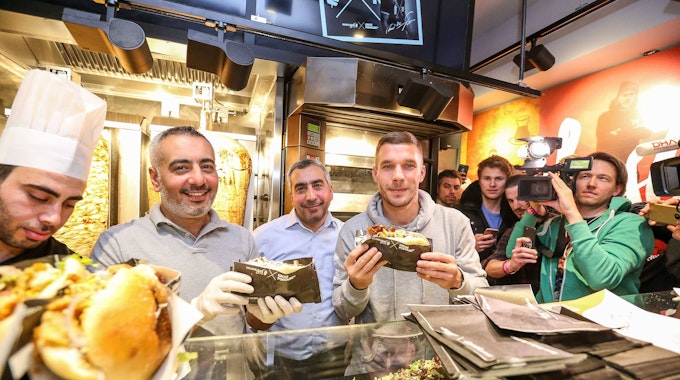 Lukas Podolski posiert mit Kollegen bei der Eröffnung eines Mangal-Döner-Ladens. Die Presse und Fans stehen im Hintergrund.