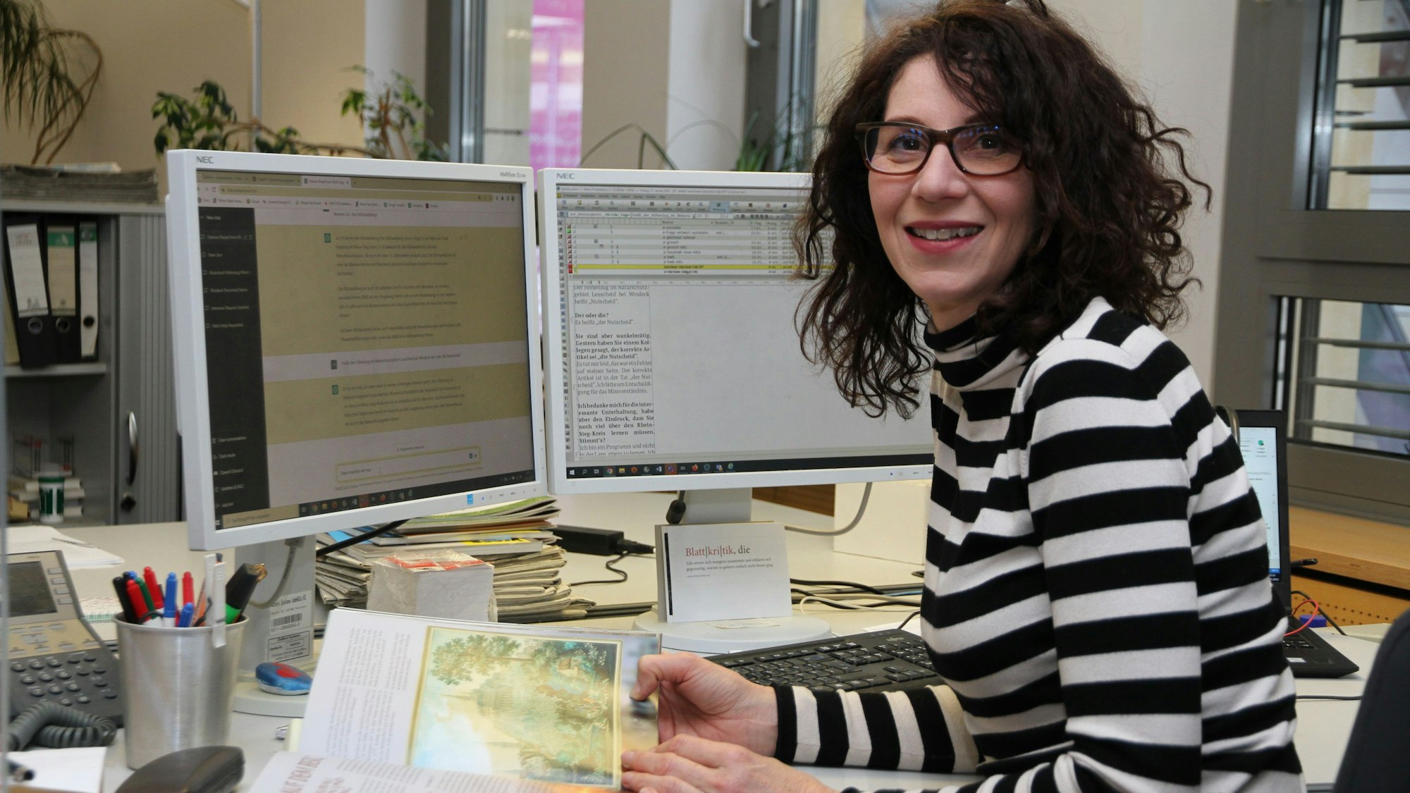 Eine Frau im gestreiften Pullover und mit Brille sitz am Schreibtisch mit einer Tastatur und zwei Bildschirmen und blättert in einem Buch.