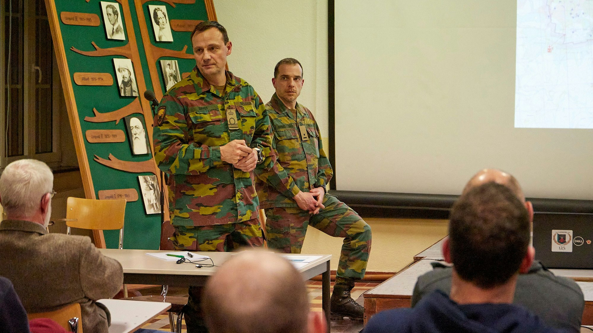 Zwei Männer in Militär-Uniformen sprechen vor einer Versammlung in einem Saal.