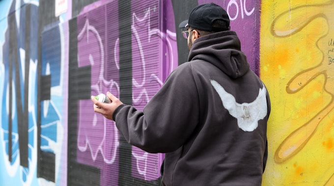 Der 22-jährige Amaru steht vor einer mit bunten Graffiti besprühten Mauer und hält eine Spraydose in der Hand.