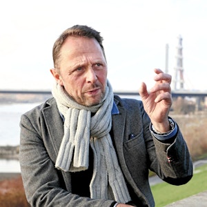 Leverkusens Oberbürgermeister Uwe Richrath, im Hintergrund sind die Rheinbrücke und der Rhein zu sehen. Richrath trägt einen Schal und gestikuliert.