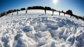 Eine Reihe Schüler fährt in der Ferne Ski, der Schnee türmt sich hoch auf, die Sonne strahlt.