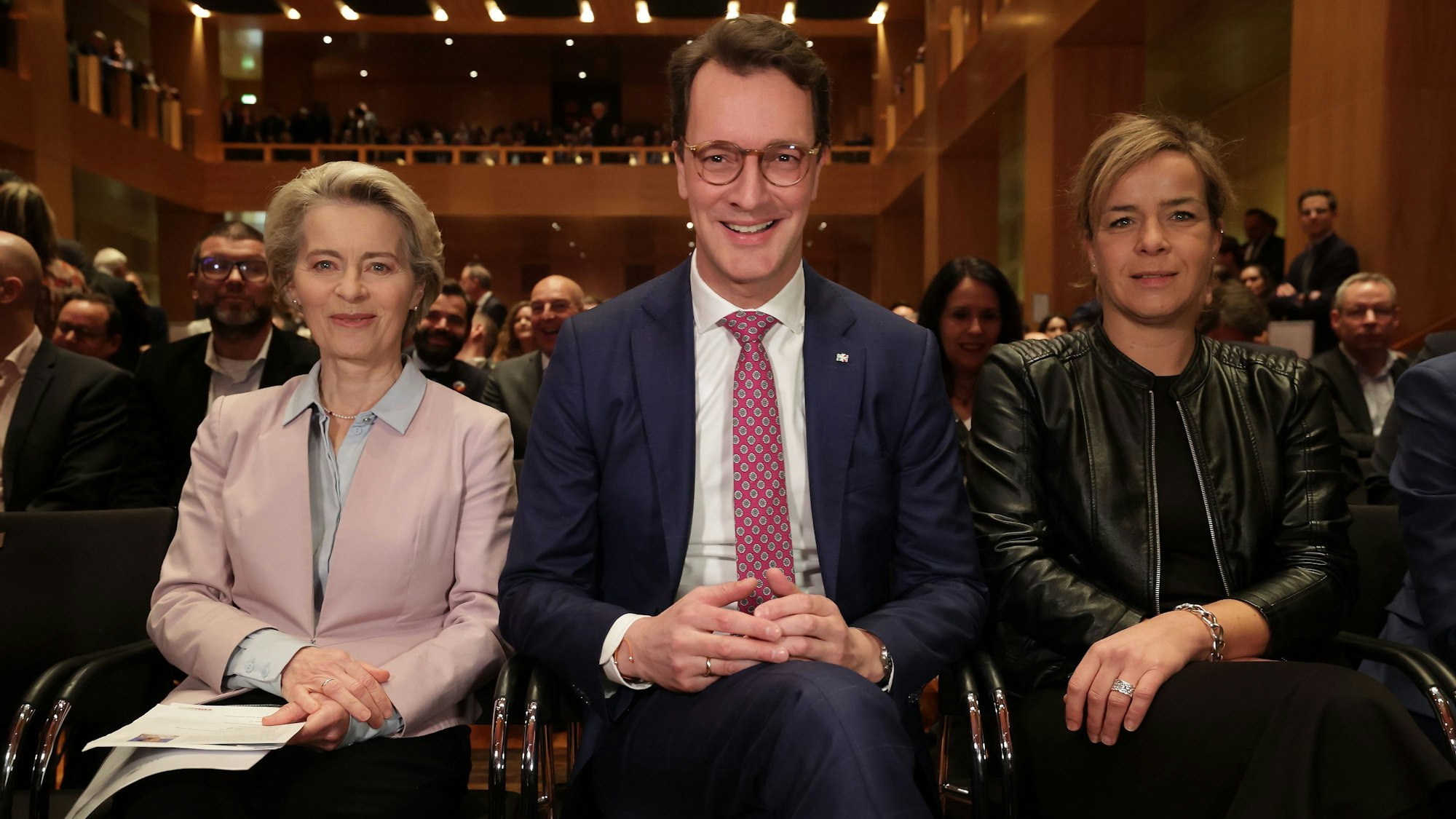 Ursula von der Leyen sitzt neben Hendrik Wüst und Ministerin Mona Neubaur. Im Hintergrund ist der mit Gästen gefüllte Saal des Kunstpalastes zu sehen.