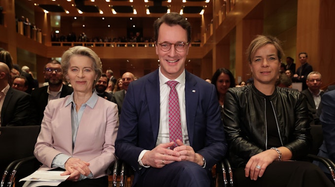 Ursula von der Leyen sitzt neben Hendrik Wüst und Ministerin Mona Neubaur. Im Hintergrund ist der mit Gästen gefüllte Saal des Kunstpalastes zu sehen.&nbsp;