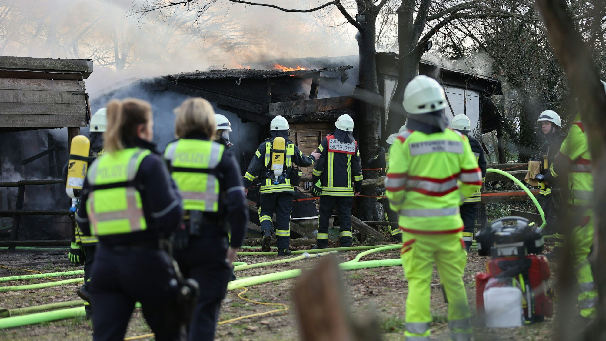 Feuerwehrleute, Sanitäter und Polizeibeamte stehen während eines Brands an einer Scheune im Kölner Stadtteil Rodenkirchen am Samstag, 28. Januar 2023. Bei dem Feuer ist eine Person tot aufgefunden worden.