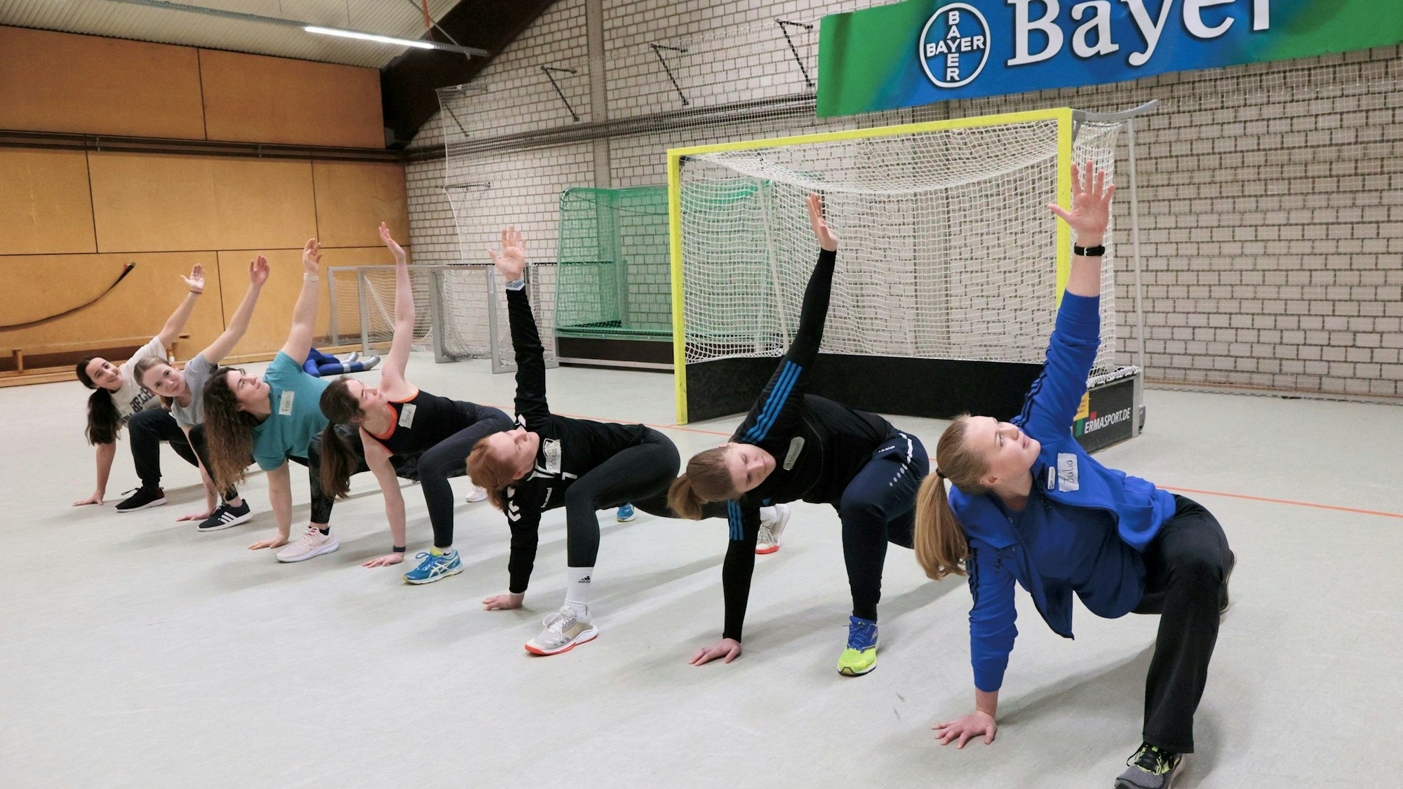 Sieben junge Frauen machen in einer Reihe in einer Sporthalle Sportübungen.