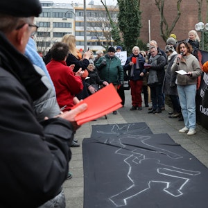 Etwa 70 Menschen kamen zu der Kundgebung vor dem Erzbischöflichen Palais in Köln. Die Teilnehmenden halten rote Karten und Transparente. Auf dem Boden ist ein schwarzes Tuch ausgebreitet, auf dem mit Kreide die Umrisse liegender Menschen nachgezeichnet sind.