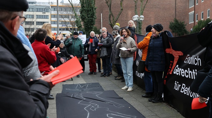 Etwa 70 Menschen kamen zu der Kundgebung vor dem Erzbischöflichen Palais in Köln. Die Teilnehmenden halten rote Karten und Transparente. Auf dem Boden ist ein schwarzes Tuch ausgebreitet, auf dem mit Kreide die Umrisse liegender Menschen nachgezeichnet sind.