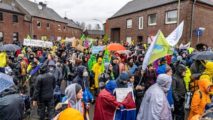 Das Bild zeigt Menschen bei einer Demo gegen den Abriss des Braunkohle Dorf Lützerath im Ort Keyenberg. Sie schwenken Fahnen und halten Schilder hoch, viele tragen Regenschutz und Schirme - das Wetter ist trüb.