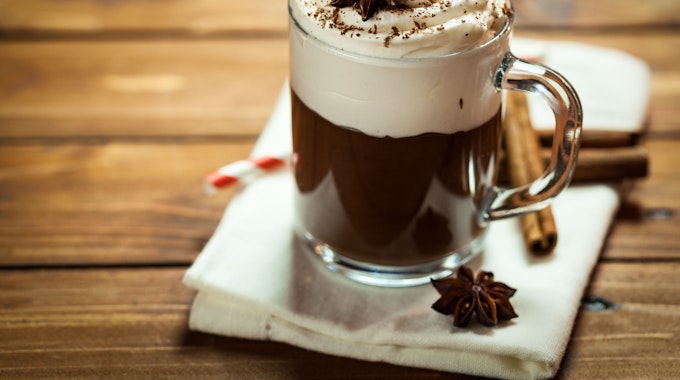 Heiße Schokolade mit Sahne, Zimt und Kakaopulver, neben der Tasse liegen dekorative Zimtstangen