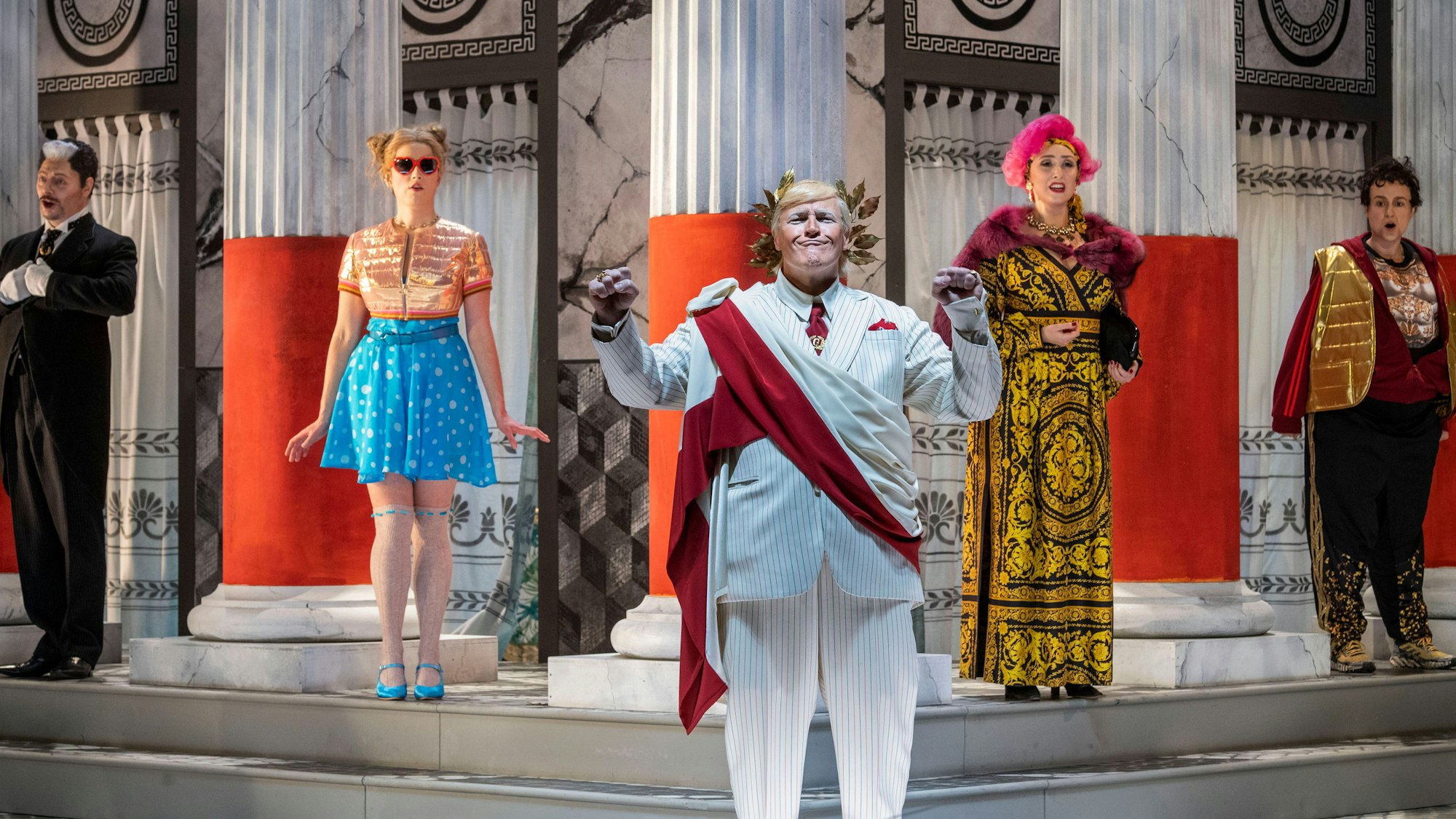 Die Bühne zeigt Säulen, Marmorwände und antike Muster. Kaiser Claudius wird als Trump inszeniert, hat einen weißen Anzug mit roter Schärpe an, aber einen goldenen Lorbeerkranz auf dem Haupt.