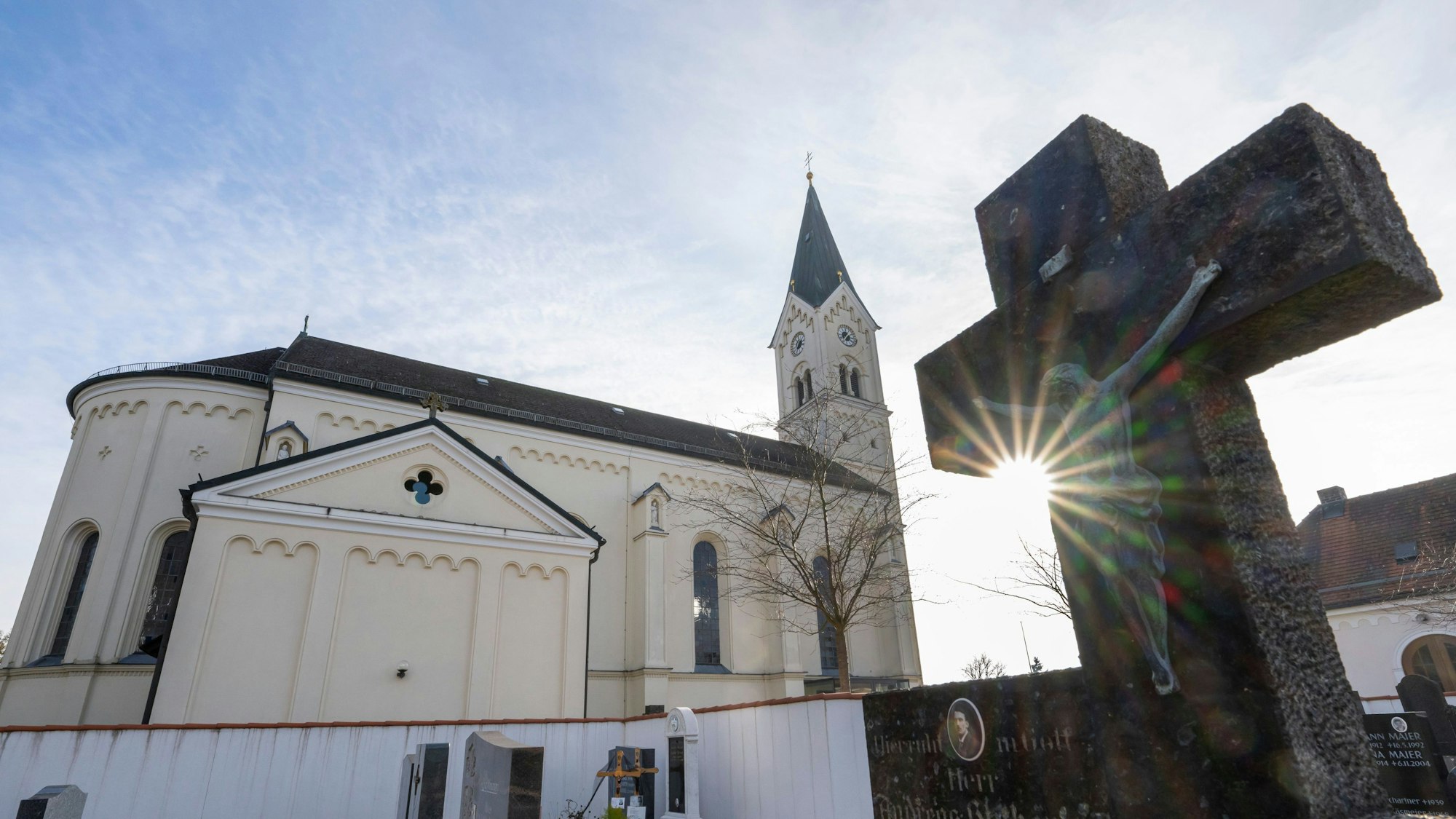 Vor dem weißen Kirchengebäude St. Nikolaus in Garching an der Alz wird ein Kreuz von der Sonne angestrahlt.