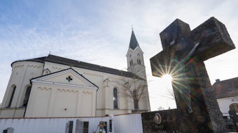 Vor dem weißen Kirchengebäude St. Nikolaus in Garching an der Alz wird ein Kreuz von der Sonne angestrahlt.