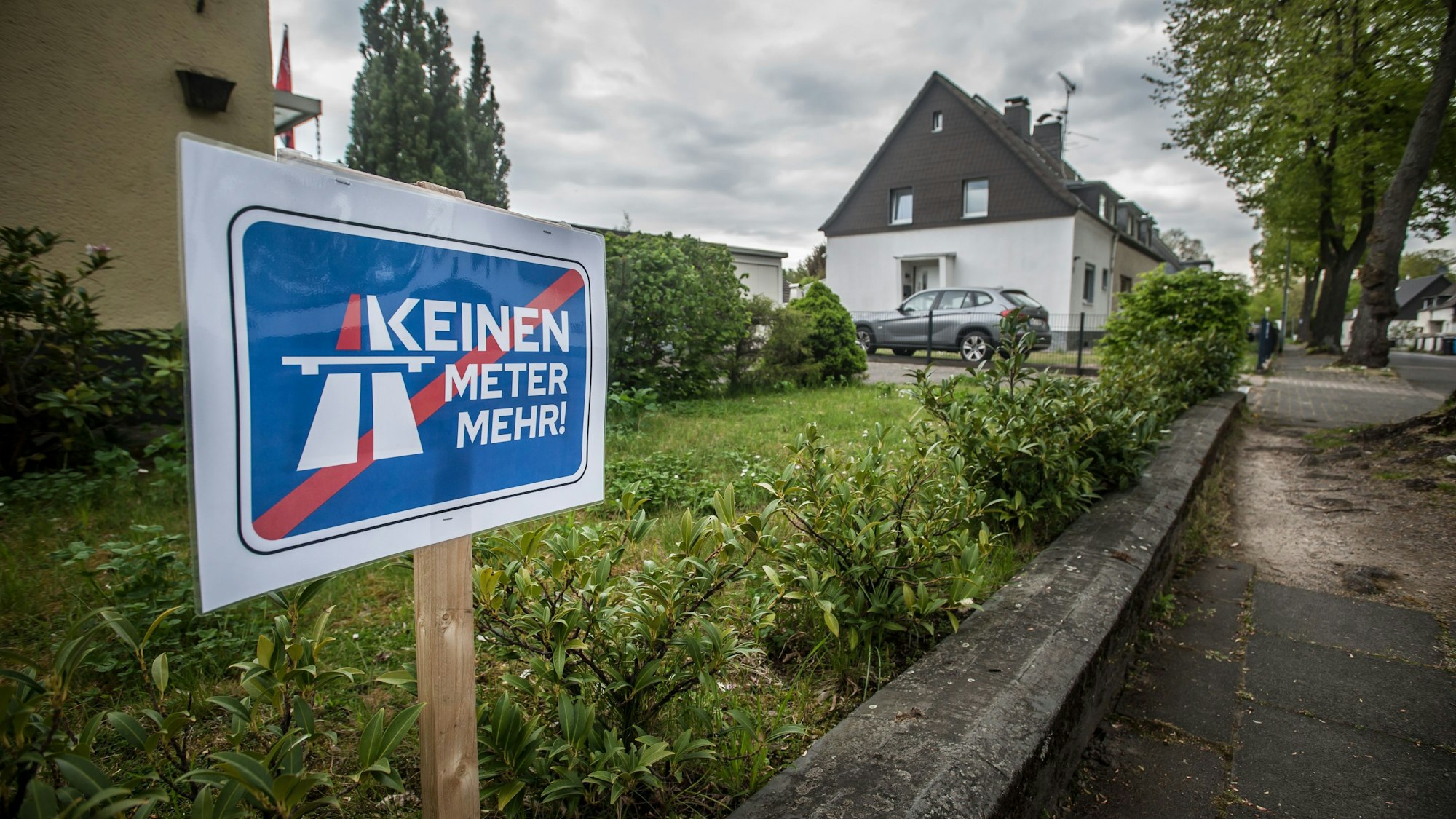 Ein Schild der Initiative „Keinen Meter mehr!“ steht in einem grünen Vorgarten, weiter hinten sind weitere Häuser und ein geparktes Auto zu sehen.