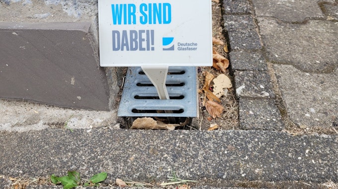 Ein Schild der Deutschen Glasfaser mit der Aufschrift „Wir sind dabei“ steckt an Gehwegrand in Kürten.