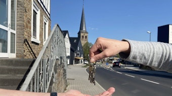 Symbolbild einer Schlüsselübergabe von Hand zu Hand, im Hintergrund eine Dorfkulisse mit Kirchturm.