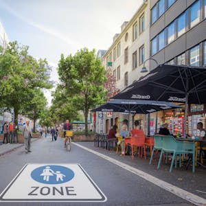 Auf einer Straße prangt riesengroß das Piktogramm für Fußgängerzone. Rechts sind Stühle und Tische eines Cafés zu sehen, Radler und Fußgänger sind auf der Straße unterwegs.&nbsp;