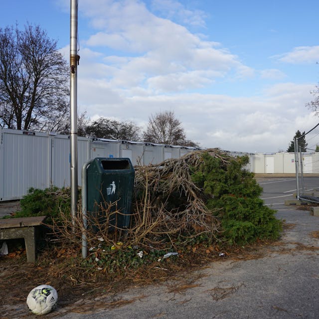 Auf einem trist aussehenden Schulhof stehen Baucontainer und Bauzäune. Dort liegen Schutt, ein alter Fußball und abgeholzte Bäume.