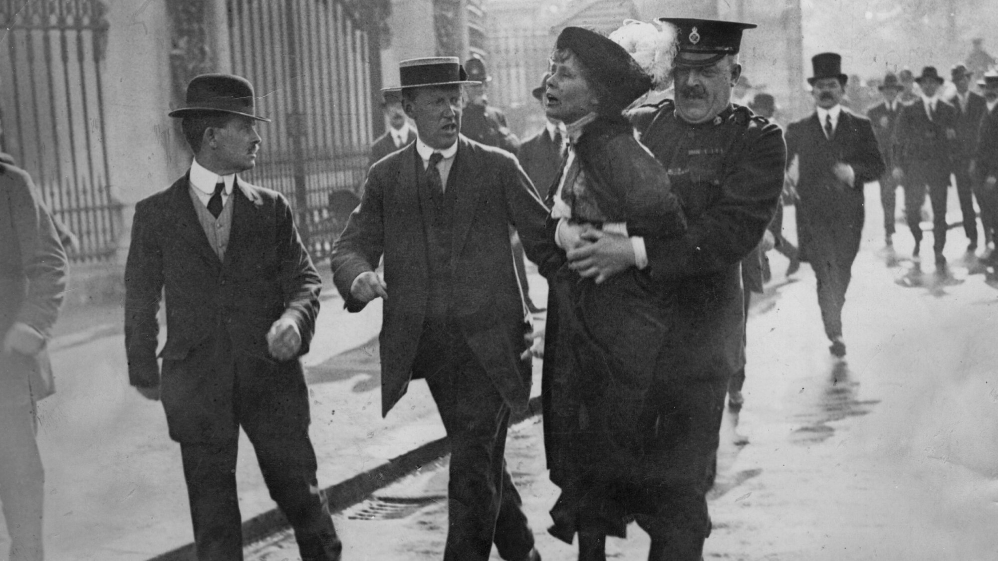 Die britische Suffragette Emmeline Pankhurst wird von einem uniformeirten Mann weggetragen.